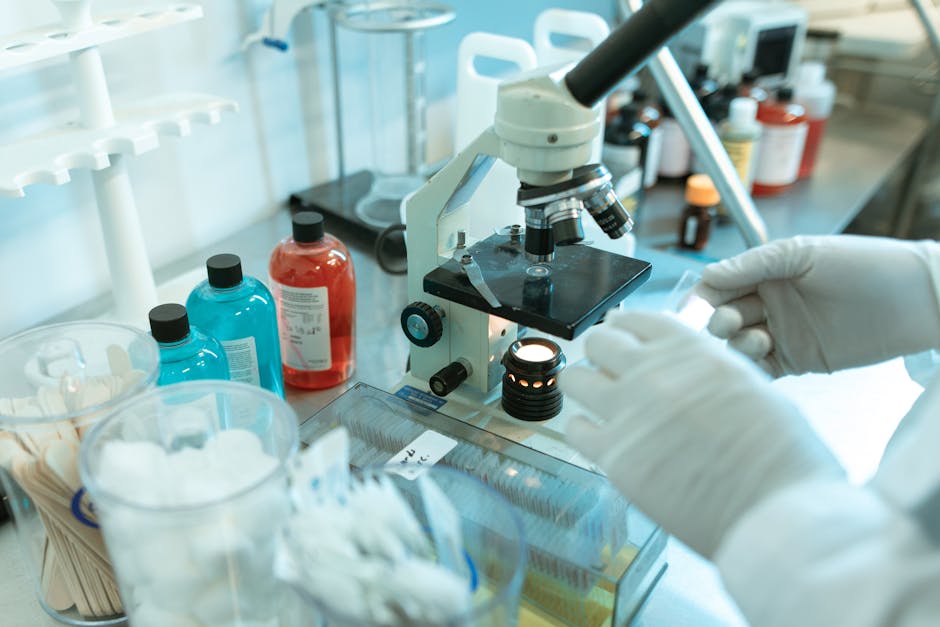 Scientist using microscope in a lab surrounded by chemicals and equipment.